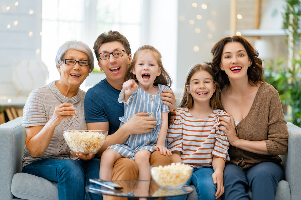 Χαρούμενη οικογένεια βλέποντας τηλεόραση με ποπ κορν στο σπίτι. Γιαγιά, μητέρα, πατέρας και κόρες περνούν χρόνο μαζί.. - Φωτογραφία, εικόνα