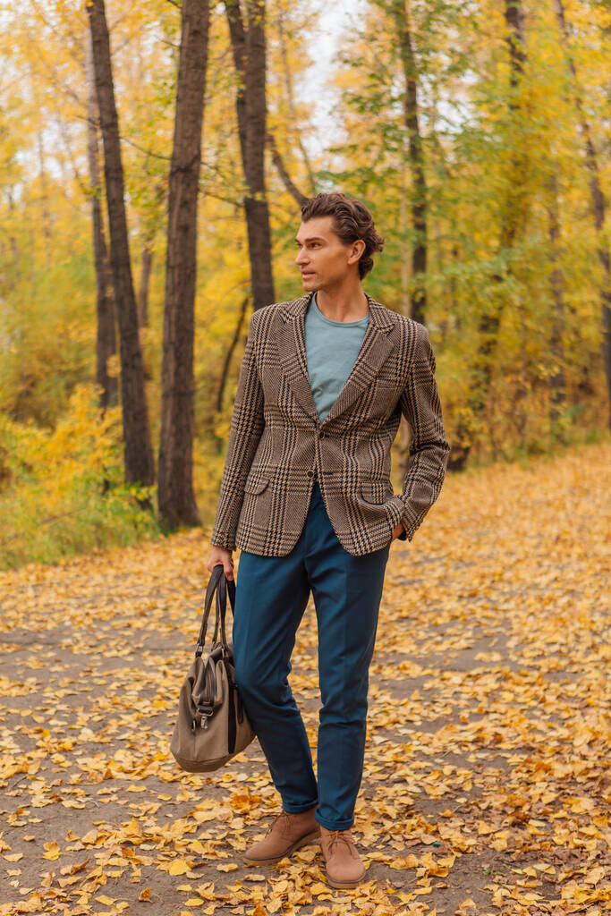 Hombre guapo alto vestido con una chaqueta marrón caminando con una bolsa en el callejón de otoño - Foto, Imagen