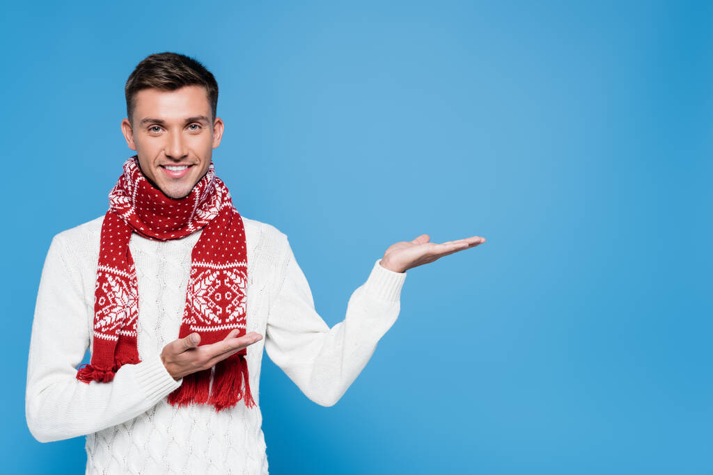 Smiling young adult man in sweater and scarf pointing with hands isolated on blue - Photo, Image