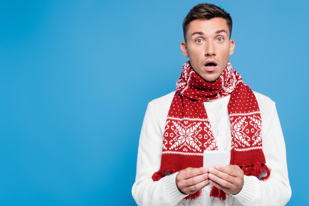 Shocked young adult man with open mouth, wearing scarf and sweater, holding smartphone, while looking at camera isolated on blue - Photo, Image