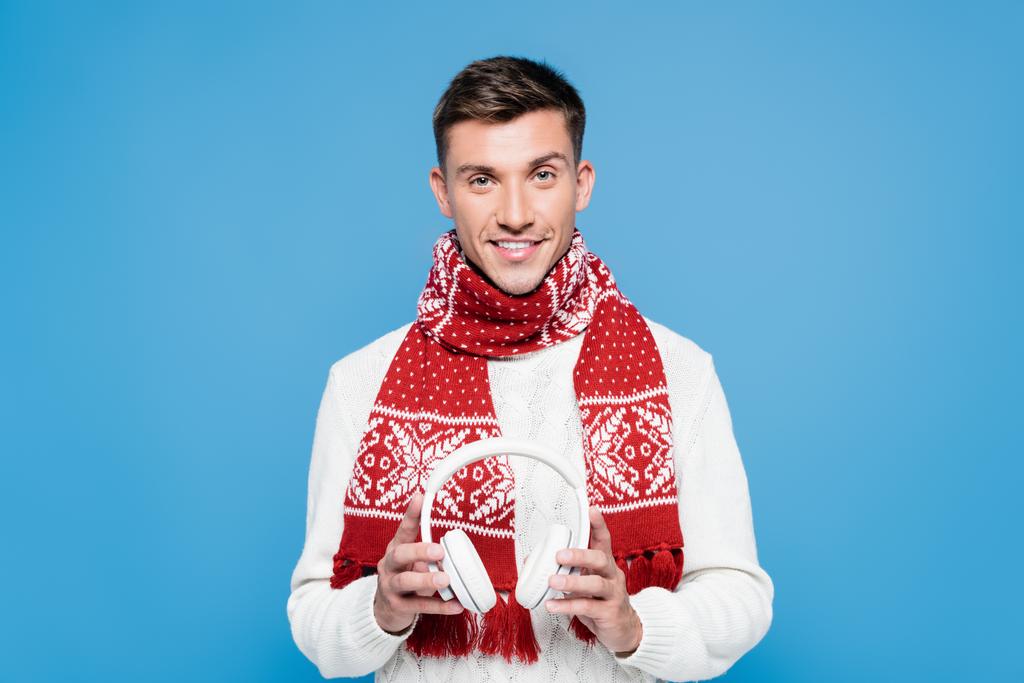 Усміхнений чоловік, одягнений светр і шарф, тримає білі бездротові навушники ізольовані на синьому
 - Фото, зображення