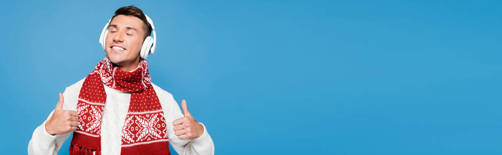 Счастливый человек с закрытыми глазами, показывающий большие пальцы вверх, в то время как носить беспроводные наушники изолированы на синий, баннер - Фото, изображение