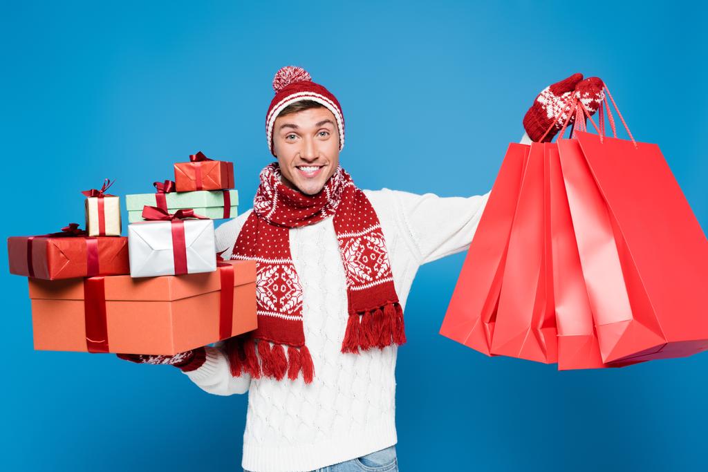 Sıcak giysili mutlu bir adamın elinde bir sürü hediye kutusu ve mavi üzerine izole edilmiş kırmızı kağıt torbalarla önü. - Fotoğraf, Görsel