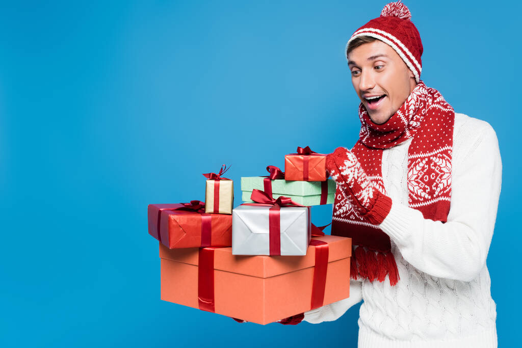 Ενθουσιασμένος άντρας με πλεκτό καπέλο και γάντια που κρατάει και κοιτάζει ένα σωρό κουτιά δώρων που είναι απομονωμένα στο μπλε - Φωτογραφία, εικόνα