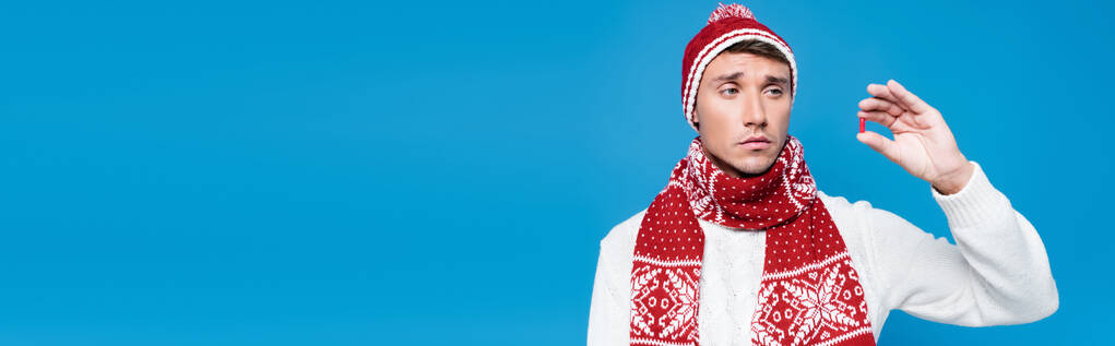 Homem doente de mau humor, vestindo roupas quentes segurando e olhando para cápsula vermelha isolada em azul, banner - Foto, Imagem