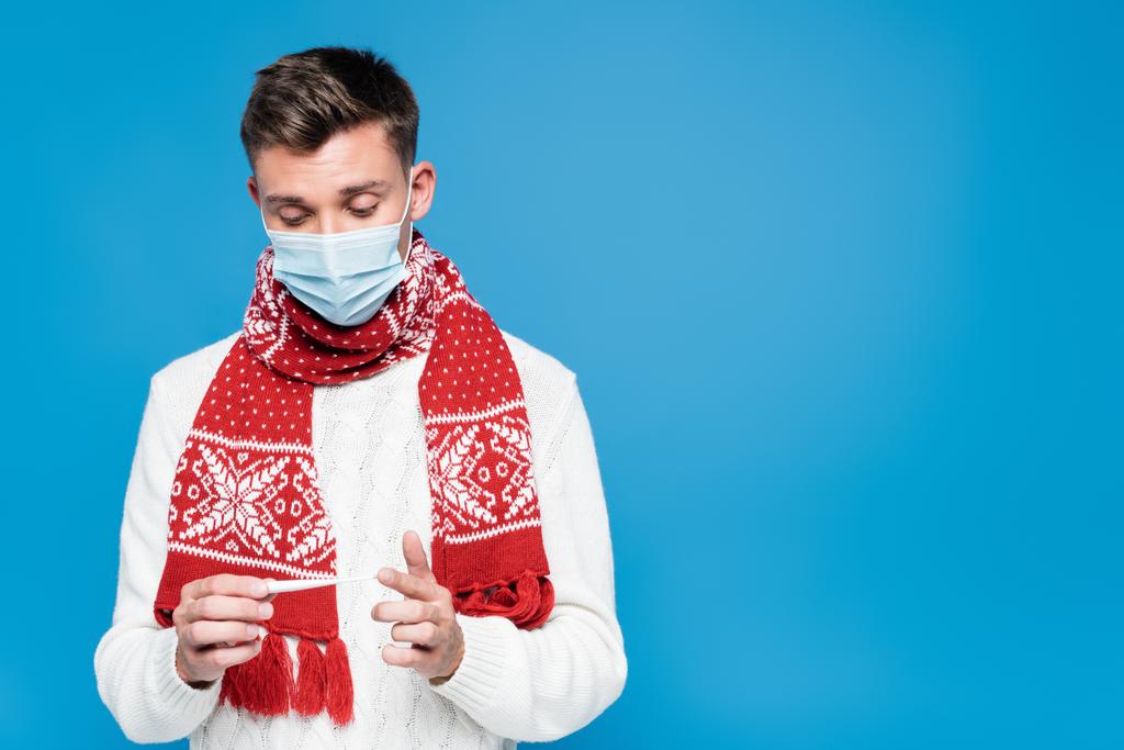 Junger erwachsener Mann mit Schal, medizinischer Maske und Blick auf Thermometer isoliert auf blau - Foto, Bild