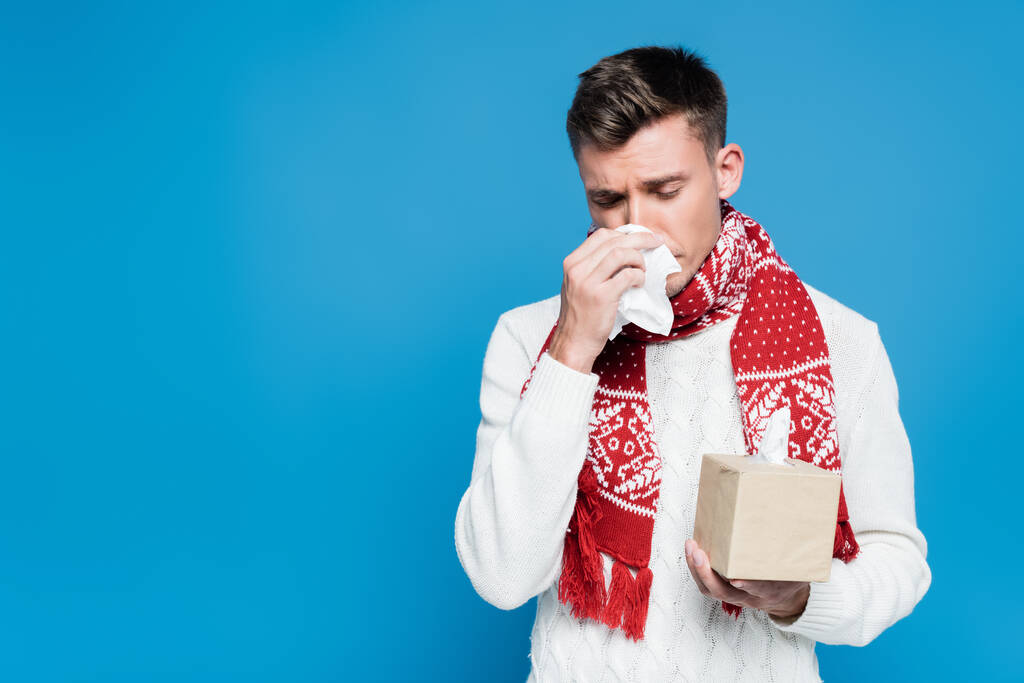 Young adult man in red scarf sneezing, while holding tissue near nose isolated on blue - Photo, Image
