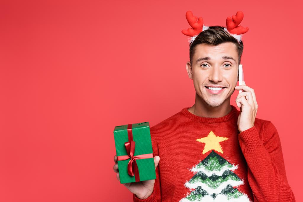 Homme souriant en pull avec pin tenant boîte cadeau et parler sur smartphone isolé sur rouge - Photo, image