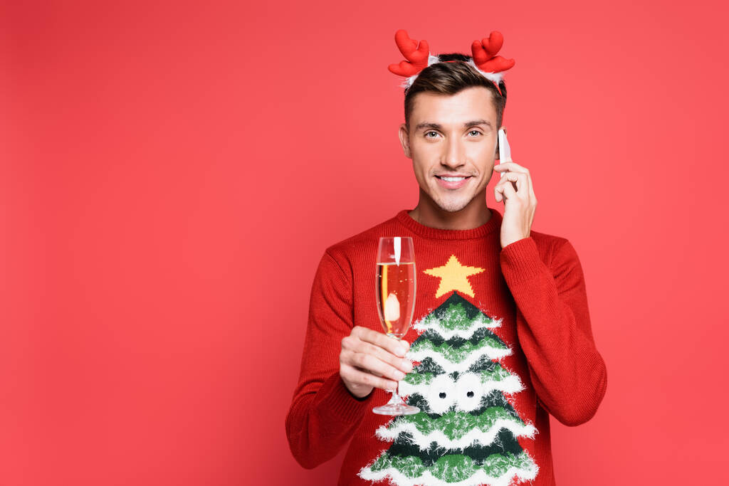 Uomo sorridente in maglione di Natale con bicchiere di champagne che parla su smartphone su sfondo rosso - Foto, immagini