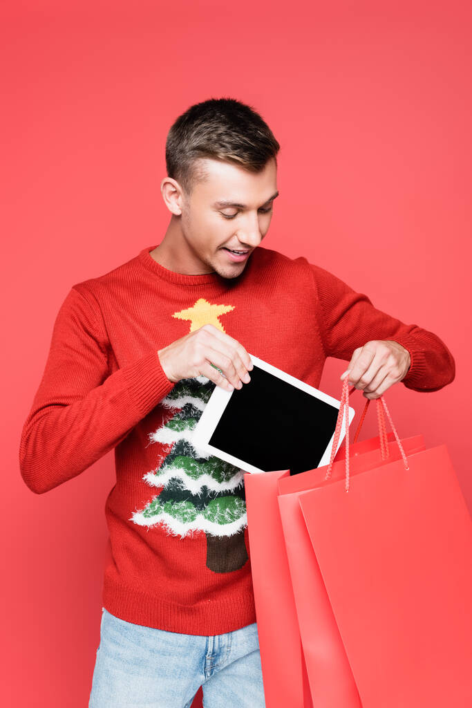 Positiver Mann im Pullover mit Kiefer steckt digitales Tablet in Einkaufstüten - Foto, Bild