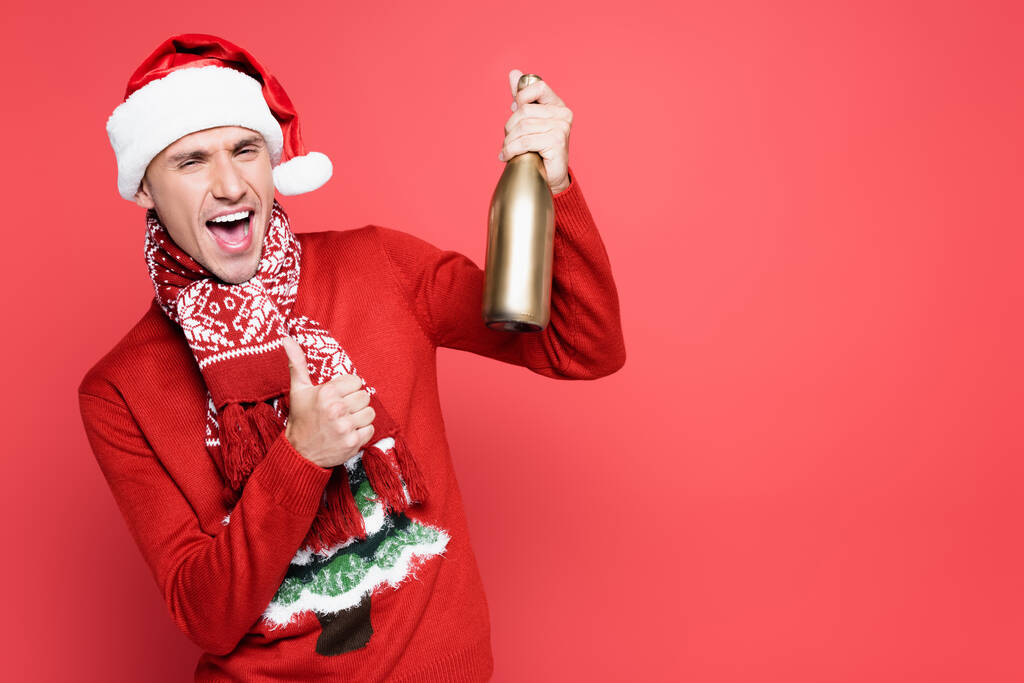 Homem excitado em Papai Noel mostrando como gesto e segurando garrafa de champanhe no fundo vermelho - Foto, Imagem