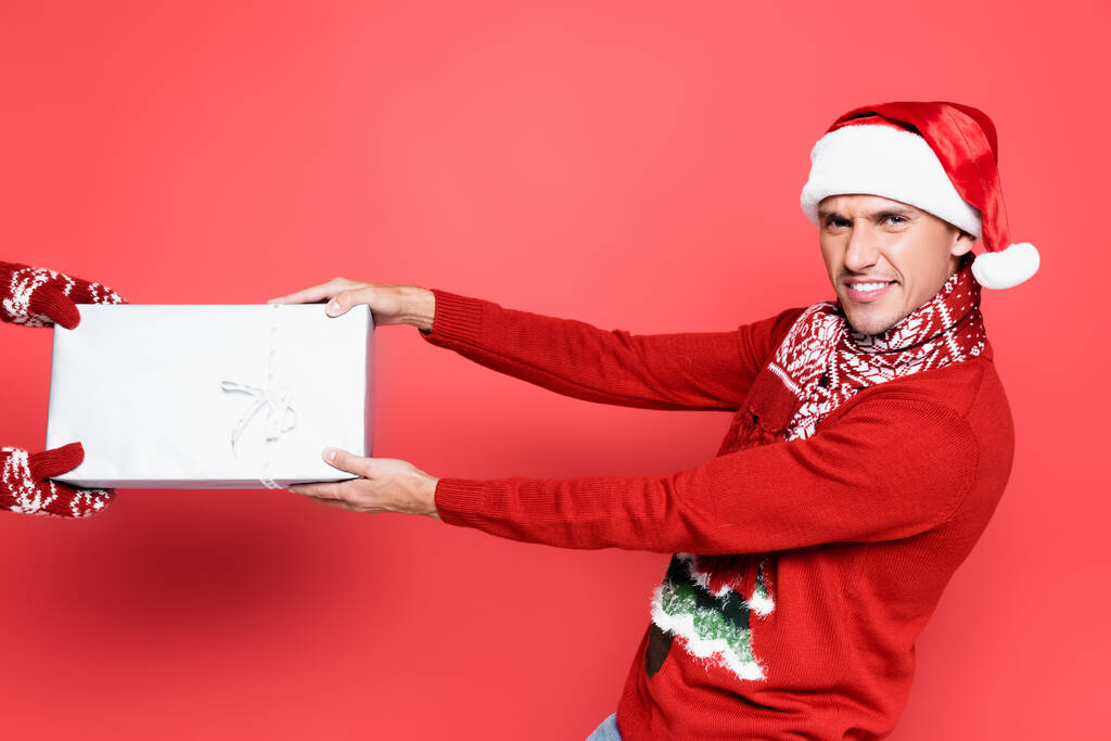 Lächelnder Mann mit Weihnachtsmütze zieht Geschenk auf rotem Hintergrund - Foto, Bild
