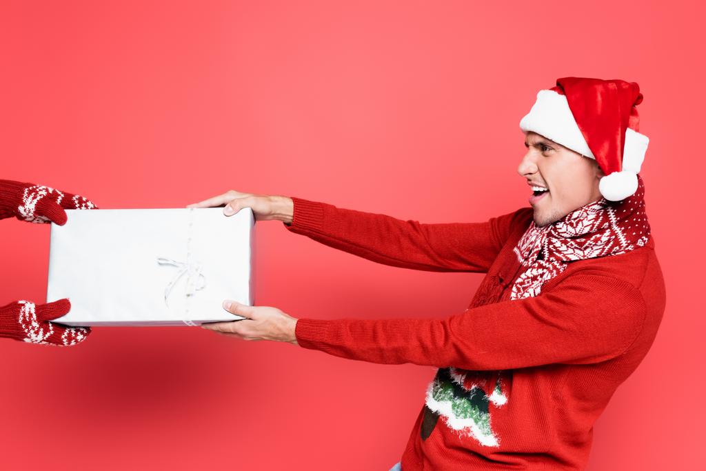 Ενθουσιασμένος άντρας με καπέλο Σάντα και μαντήλι τραβώντας κουτί δώρου σε κόκκινο φόντο - Φωτογραφία, εικόνα