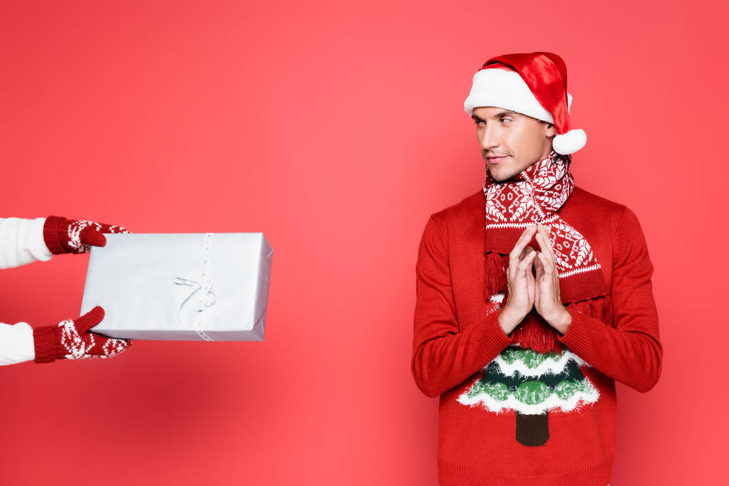 Sly man in scarf and santa hat looking at gift box on red background - Photo, Image