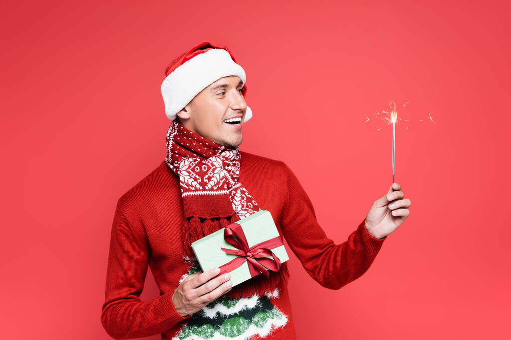 Kırmızı eşarplı, Noel Baba şapkalı, elinde maytap ve hediye kutusu olan gülümseyen bir adam. - Fotoğraf, Görsel