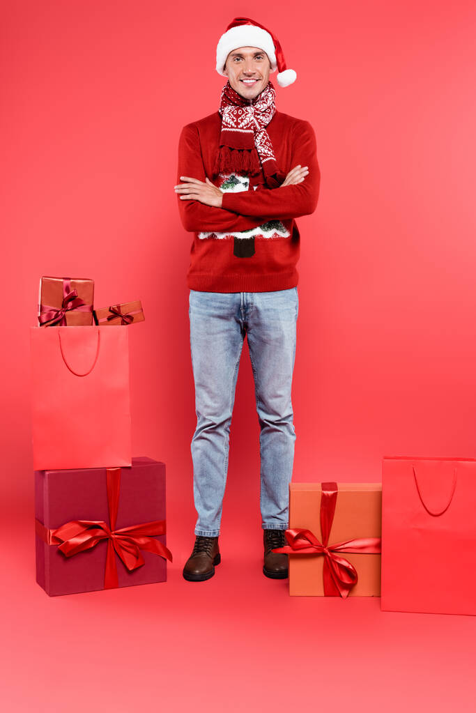 Noel Baba şapkalı gülümseyen adam kollarını kavuşturup hediye kutularının yanındaki kameraya ve kırmızı arka plandaki alışveriş çantalarına bakıyor. - Fotoğraf, Görsel