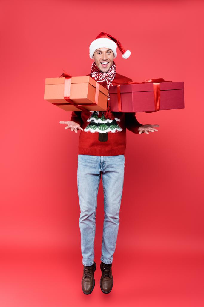 Positivo uomo in cappello di Babbo Natale saltando vicino presenta su sfondo rosso - Foto, immagini