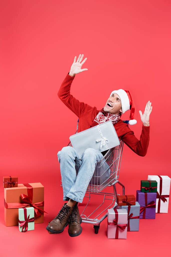 Hombre alegre en bufanda y sombrero de santa sentado en el carrito de la compra cerca de regalos sobre fondo rojo - Foto, imagen