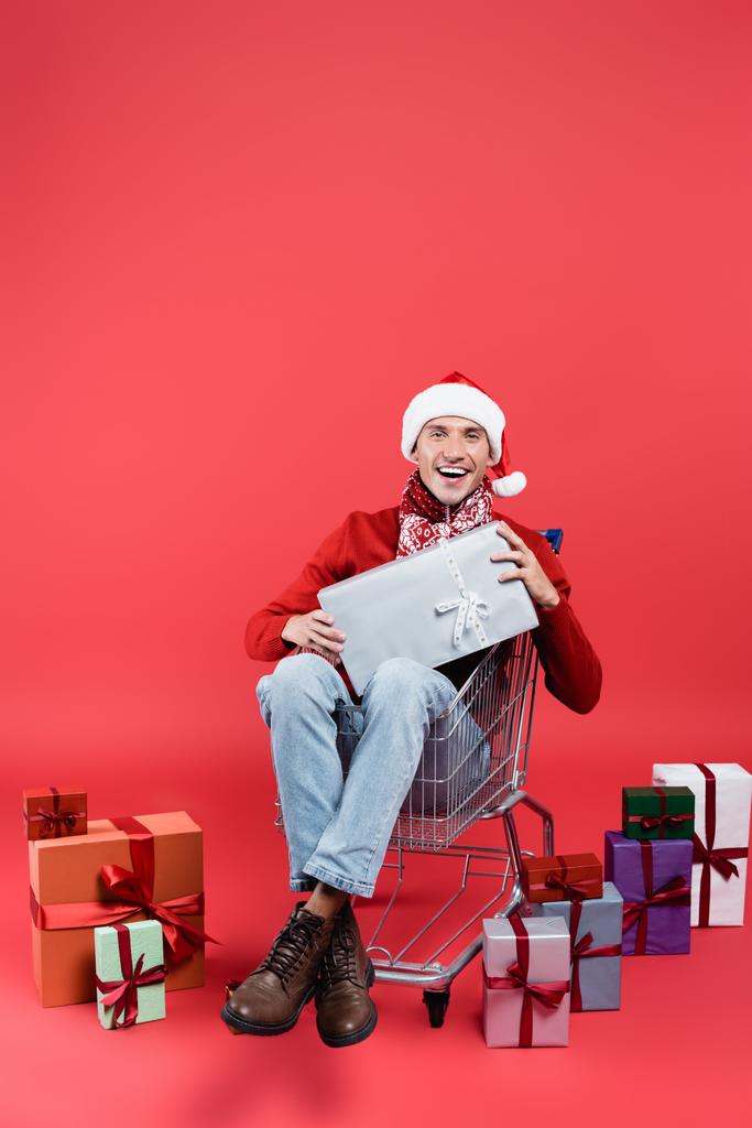 Mosolygós férfi télapó kalap nézi a kamerát, miközben kezében ajándék bevásárlókocsi piros háttér - Fotó, kép