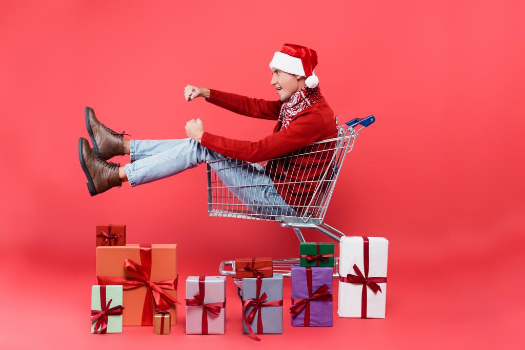 Seitenansicht eines jungen Mannes mit Weihnachtsmütze, der in einem Einkaufswagen in der Nähe von Geschenkboxen auf rotem Hintergrund sitzt - Foto, Bild
