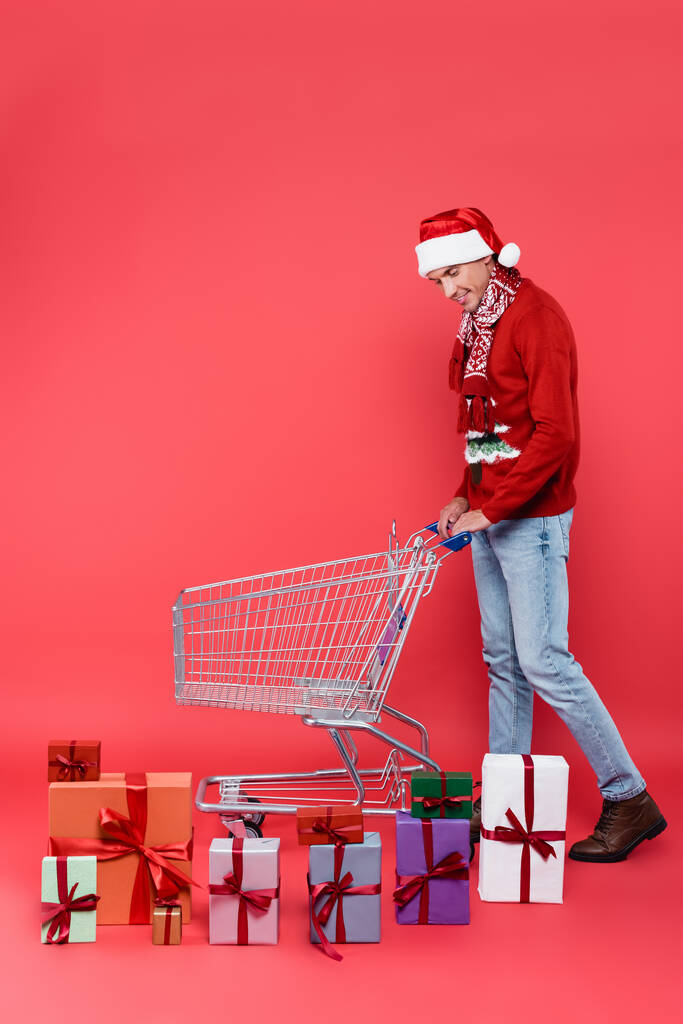 Hombre sonriente en bufanda y sombrero de santa de pie cerca de regalos y carrito de compras sobre fondo rojo - Foto, imagen