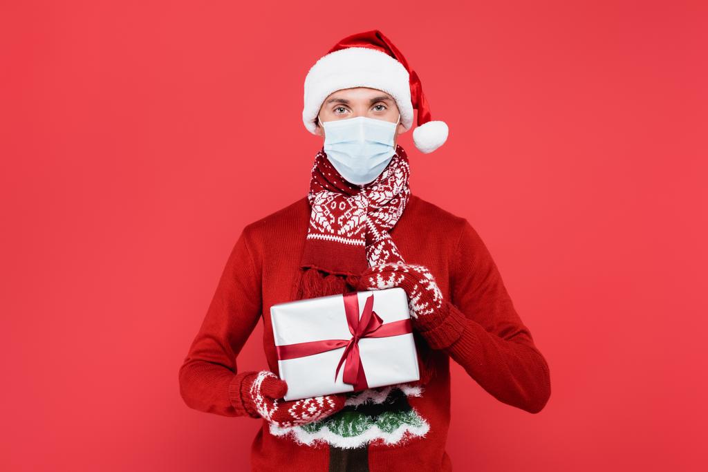 Młody mężczyzna w czapce Mikołaja, masce medycznej i rękawiczkach trzymając pudełko prezentów odizolowany na czerwono  - Zdjęcie, obraz