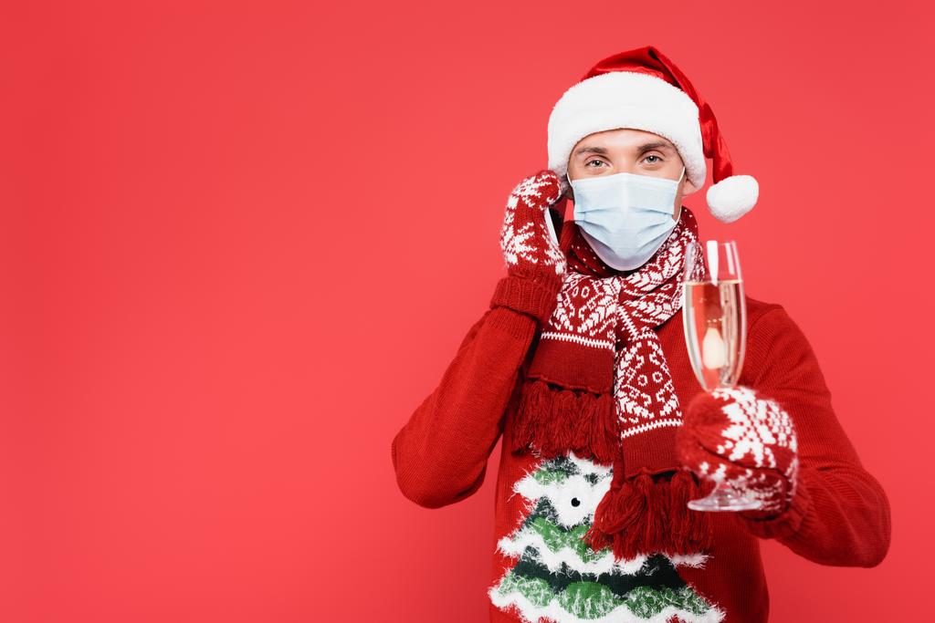 Man in santa hat and medical mask talking on smartphone while holding glass of champagne on blurred foreground isolated on red  - Photo, Image