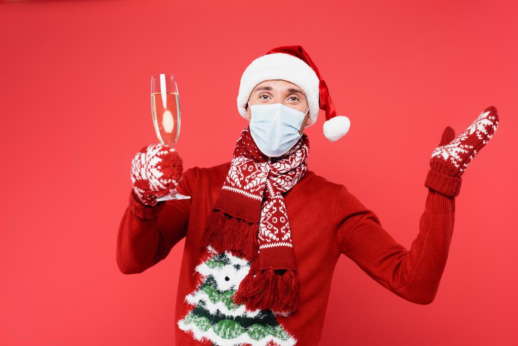 Tıbbi maskeli ve Noel Baba şapkalı bir adam kırmızı üzerine izole edilmiş şampanya bardağı tutuyor.  - Fotoğraf, Görsel
