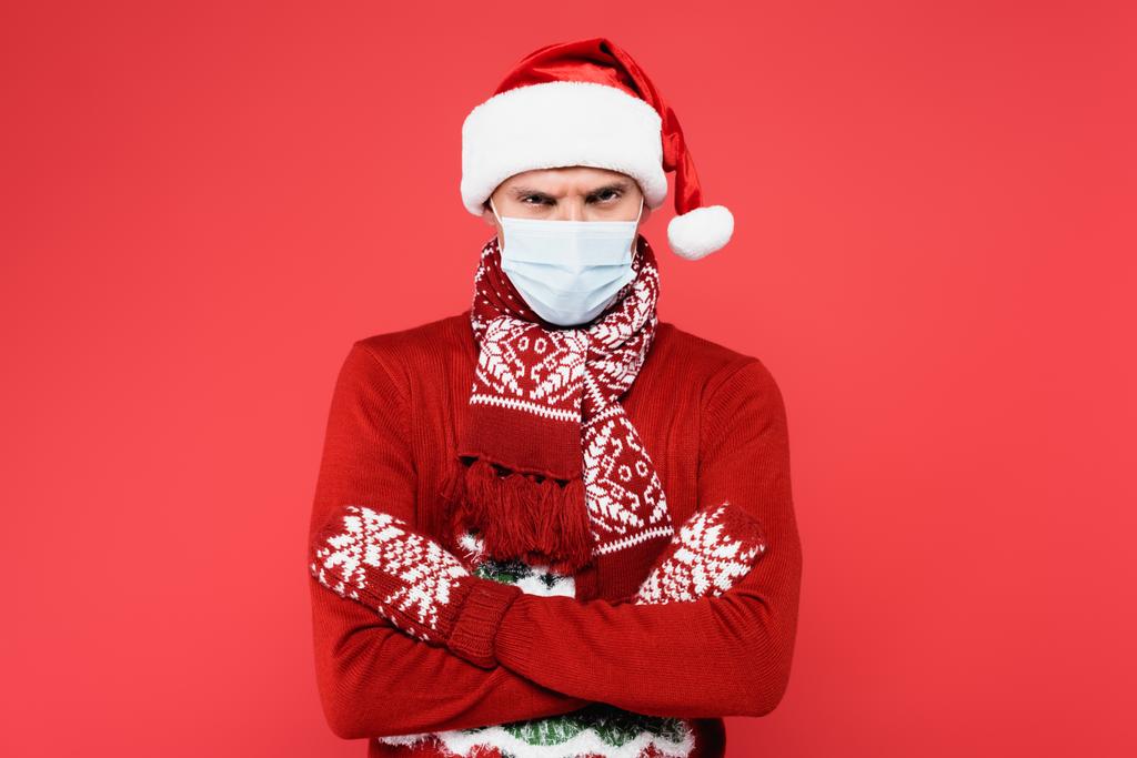 Vihainen mies lääketieteellisessä naamiossa ja jouluhattu katsellen kameran eristetty punaisella  - Valokuva, kuva