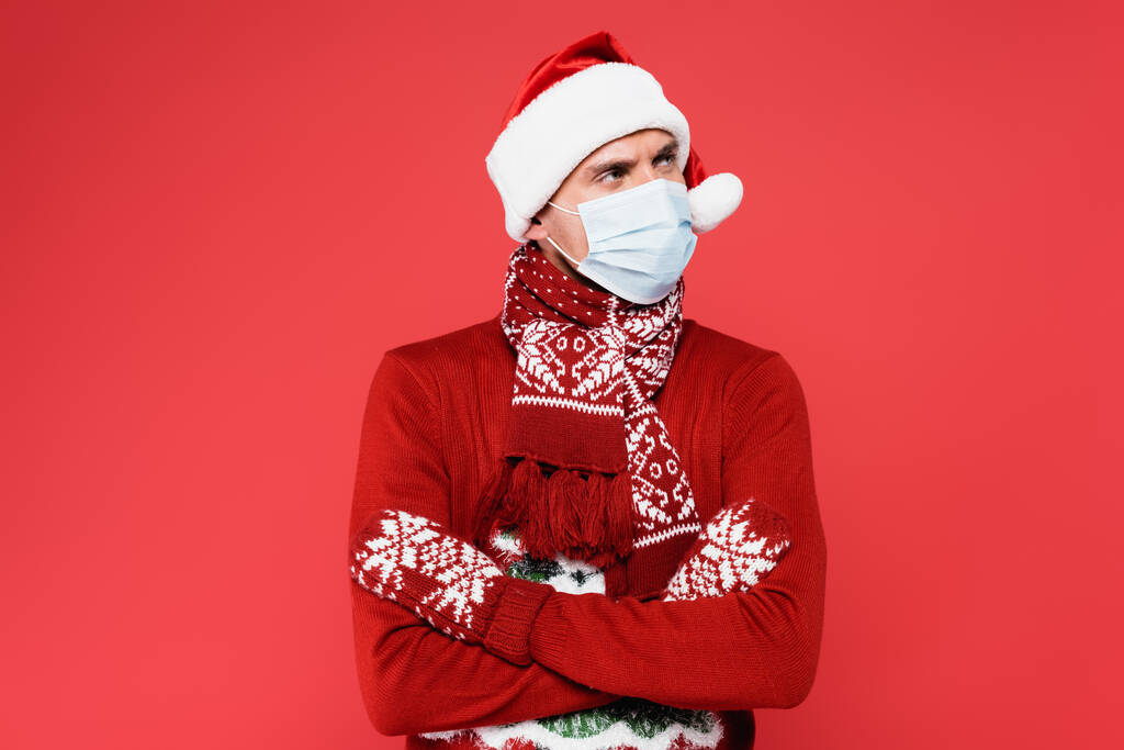 Noel Baba şapkalı, tıbbi maskeli ve eldivenli genç adam kırmızı kollarında çapraz kollarla dikiliyor.  - Fotoğraf, Görsel