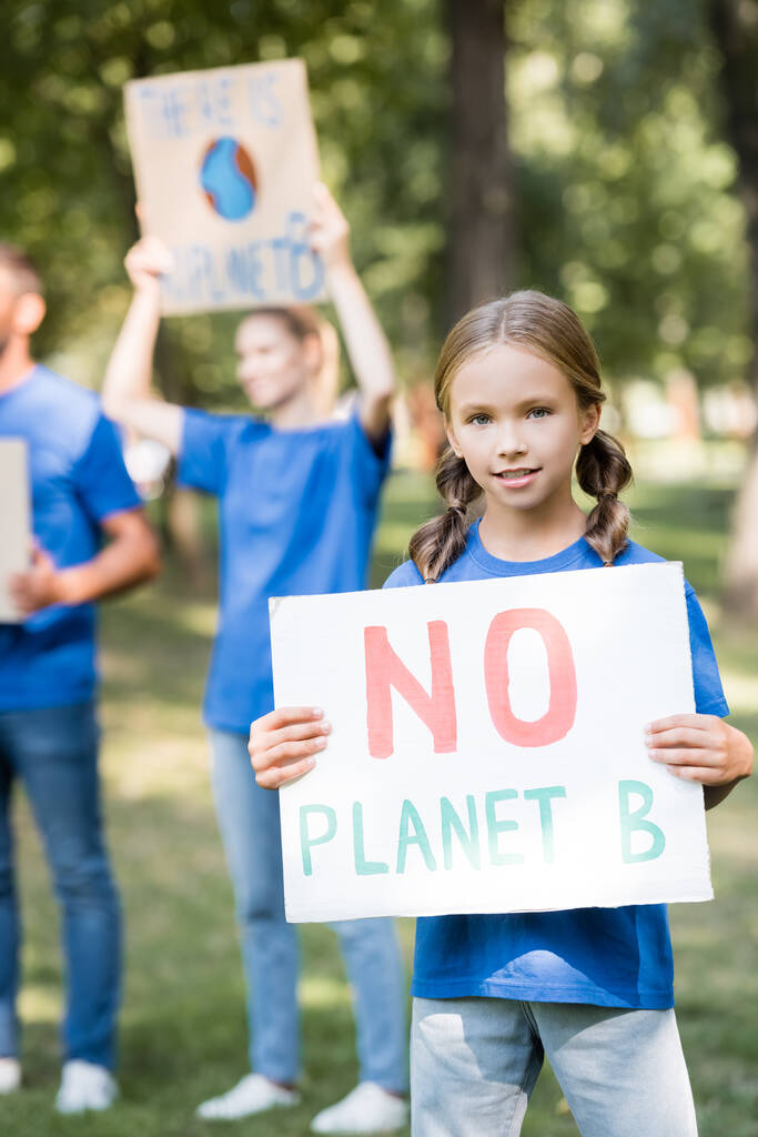 девушка держит плакат без надписи на планете Б рядом с семьей с плакатами на размытом фоне, экологическая концепция - Фото, изображение