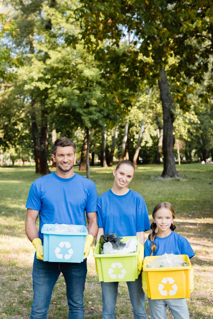 glimlachende vrijwilligers familie houden containers met recycling emblemen, vol met plastic afval, ecologie concept - Foto, afbeelding