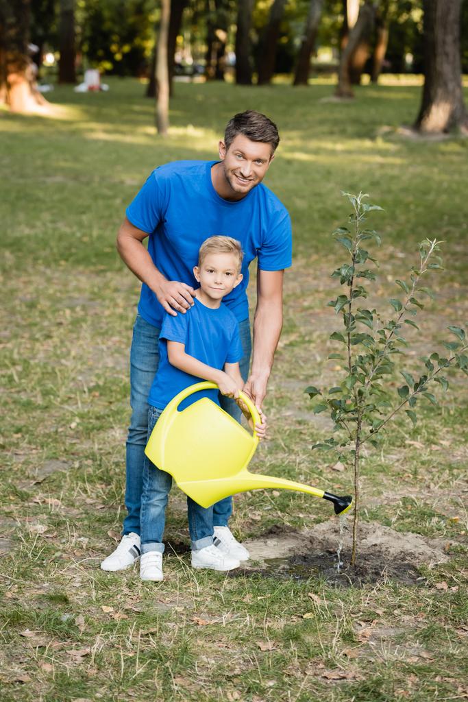 boy looking at camera while watering planted young tree near smiling father, ecology concept - Photo, Image