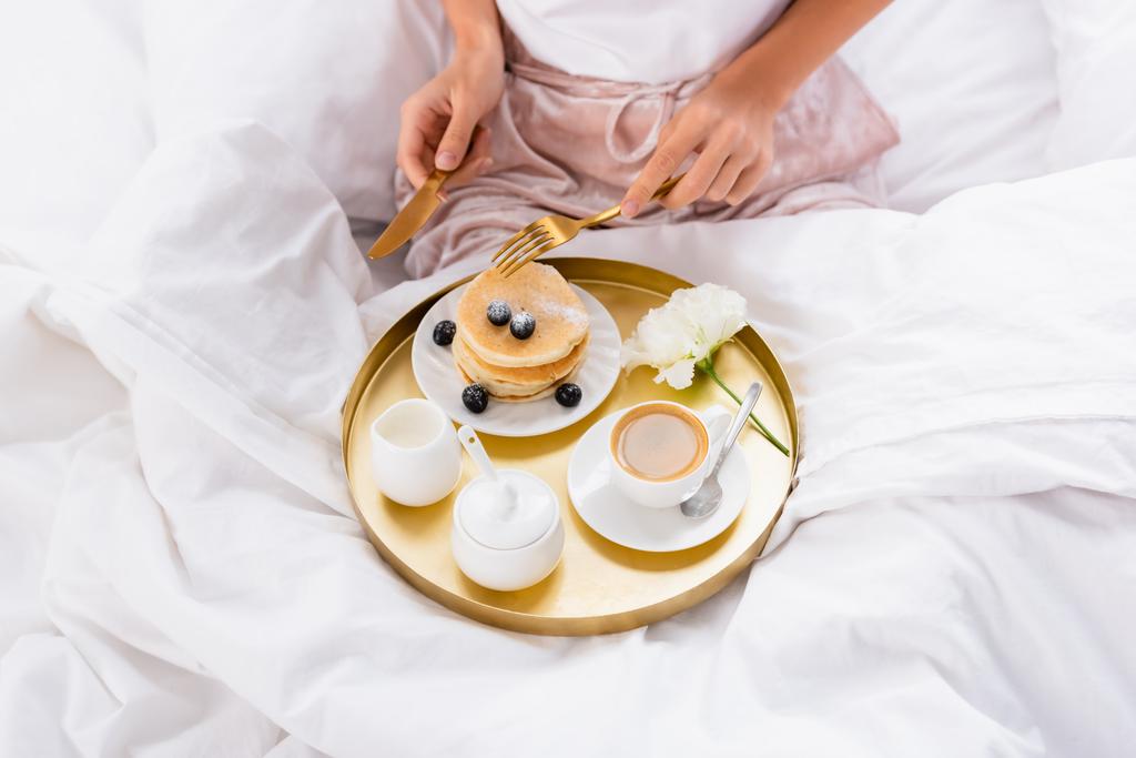 ベッドの上で朝食トレイにパンケーキ、コーヒー、カーネーションの近くに刃物を保持する女性の作物ビュー  - 写真・画像