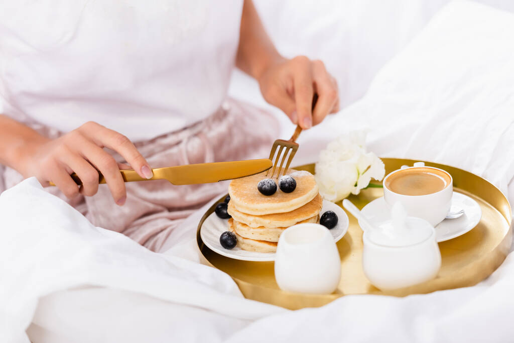 Rajatut näkymät ruokailuvälineet käsissä nainen lähellä pannukakkuja marjoja, kahvia ja neilikka aamiaisella tarjotin sängyllä  - Valokuva, kuva