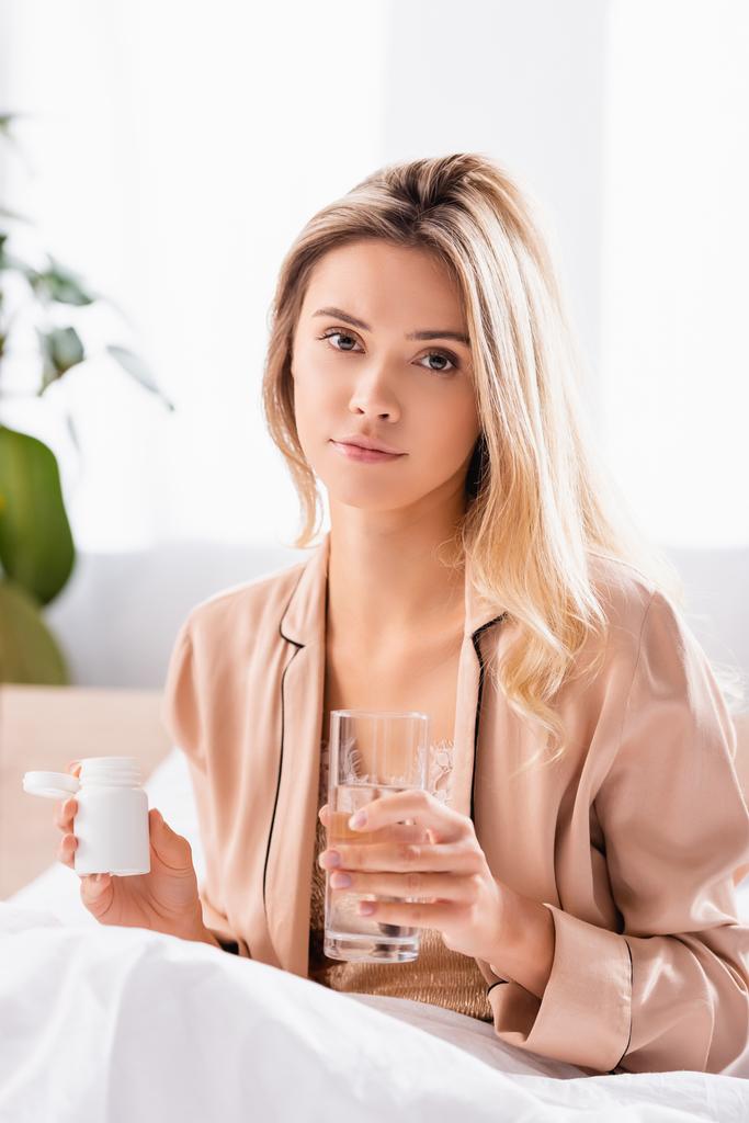 Молода жінка дивиться на камеру, тримаючи склянку води і банку з таблетками на ліжку вранці
  - Фото, зображення