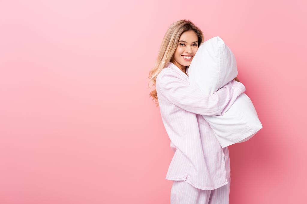 Mujer joven en pijama sonriendo a la cámara mientras sostiene la almohada sobre fondo rosa - Foto, imagen