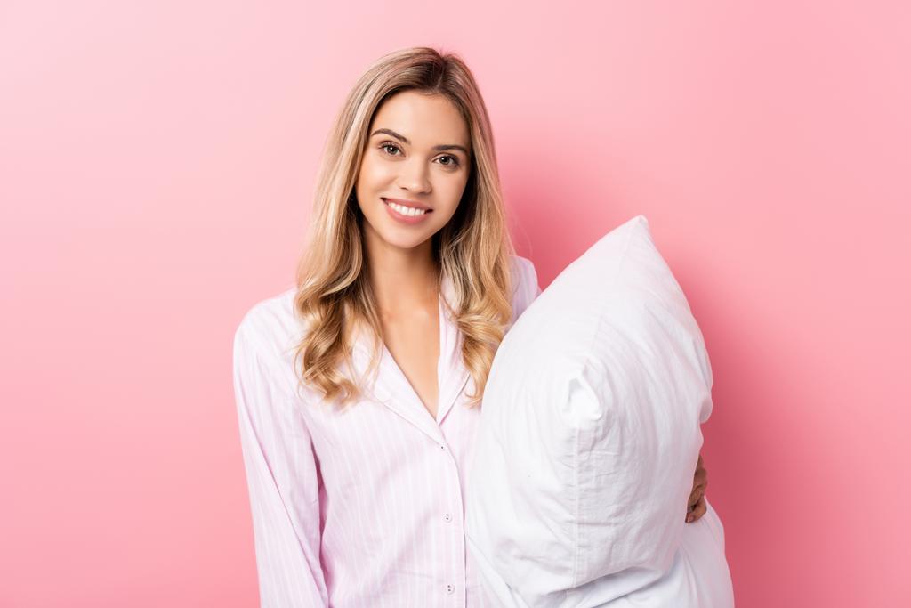 Giovane donna in pigiama sorridente alla macchina fotografica e tenendo il cuscino su sfondo rosa - Foto, immagini