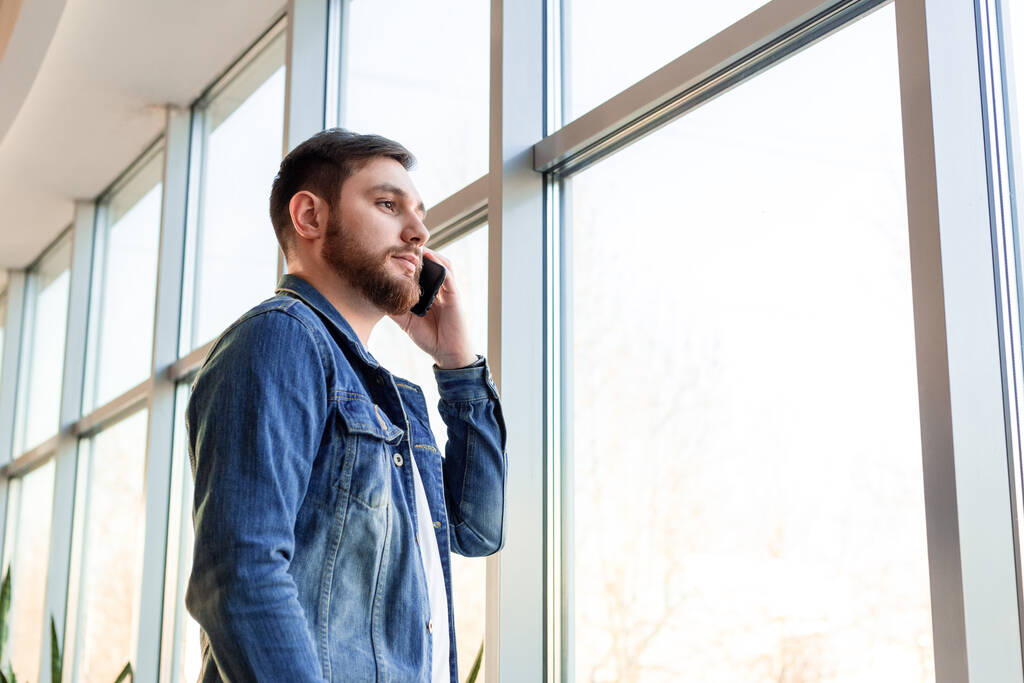 Junger Mann in lässiger Jeansjacke telefoniert drinnen in der Nähe der Fensterwand. Kluge Männer in modernen Großstadtbüros unterhalten sich mobil. Kaukasischer bärtiger Geschäftsmann - Foto, Bild