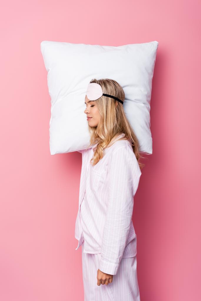 Jeune femme en pyjama et bandeau tenant un oreiller sur fond rose - Photo, image