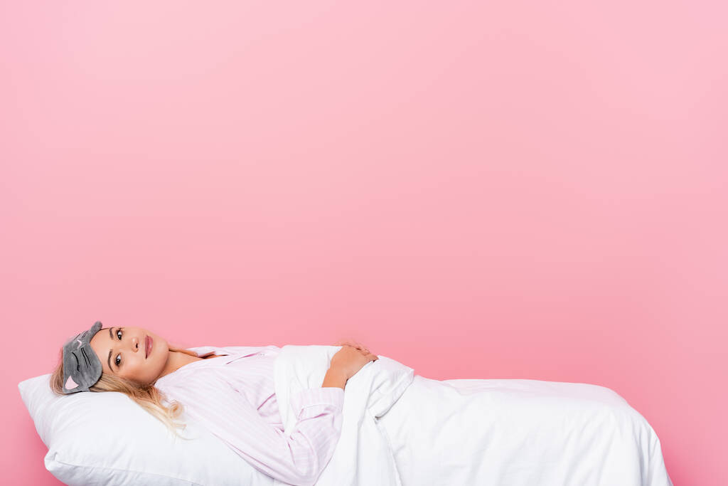 fiatal nő pizsamában és bekötött szemmel nézi a kamerát, miközben feküdt ágynemű rózsaszín háttér - Fotó, kép