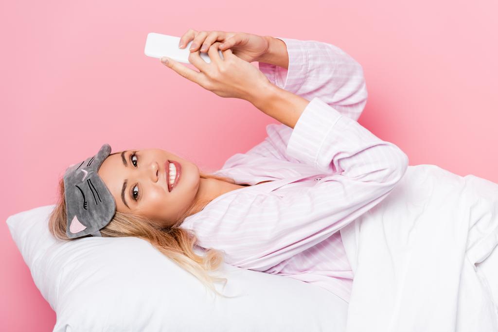 Mujer alegre en pijama y venda de ojos usando smartphone en ropa de cama sobre fondo rosa - Foto, Imagen