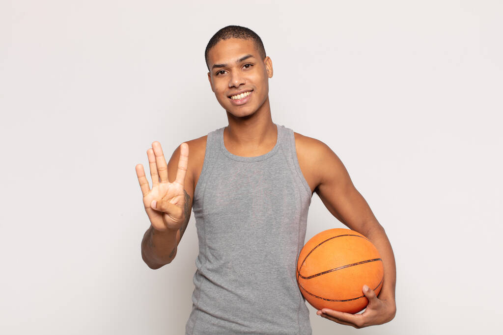 jonge zwarte man glimlachen en kijken vriendelijk, tonen nummer vier of vierde met de hand vooruit, aftellen - Foto, afbeelding