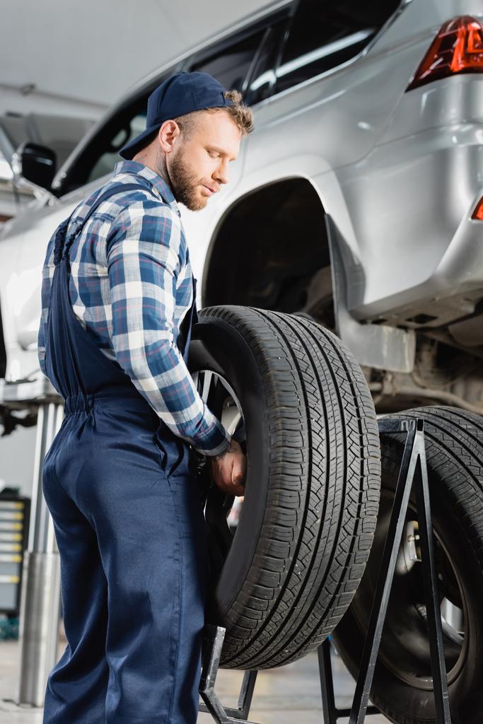 mechanic in overalls holding wheel near automobile raised on car lift on blurred background - Photo, Image