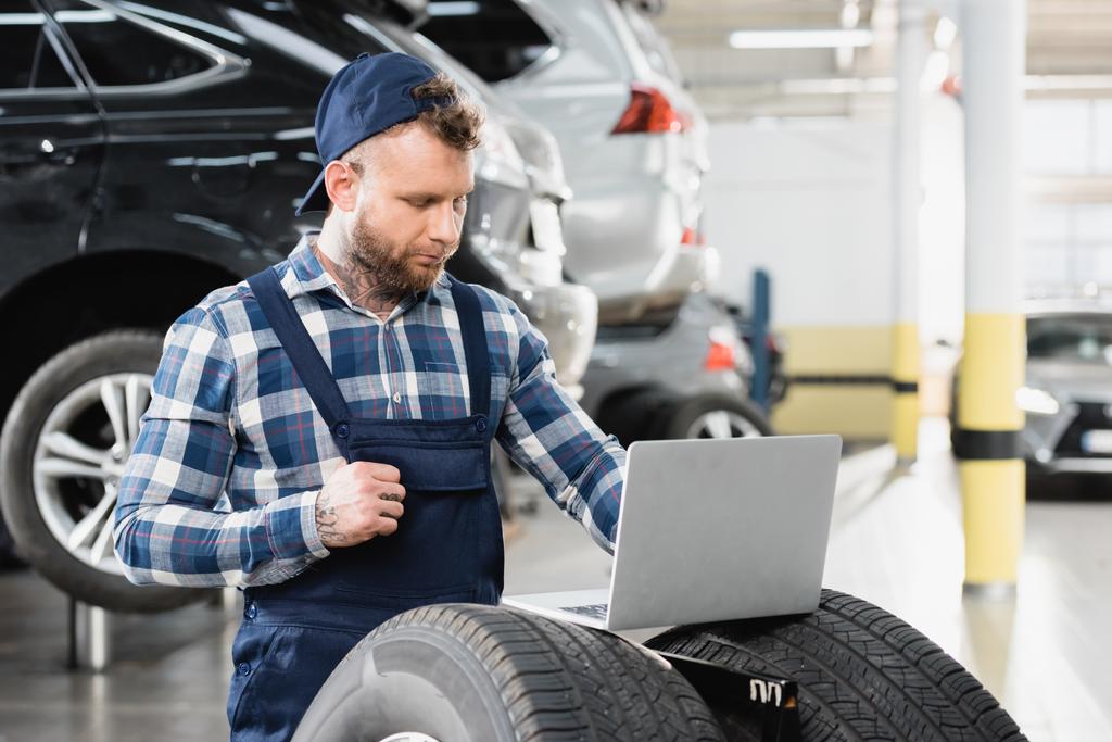young technician working on laptop near cars in workshop on blurred background - Photo, Image