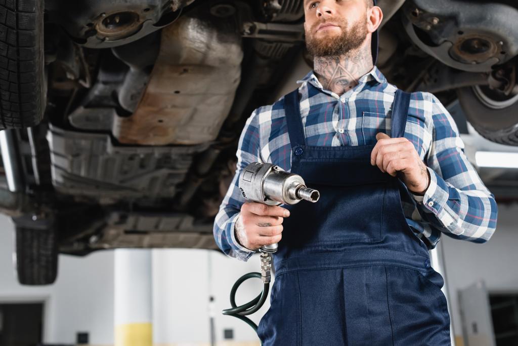 low angle view of technician holding pneumatic wrench near raised car on blurred background - Photo, Image