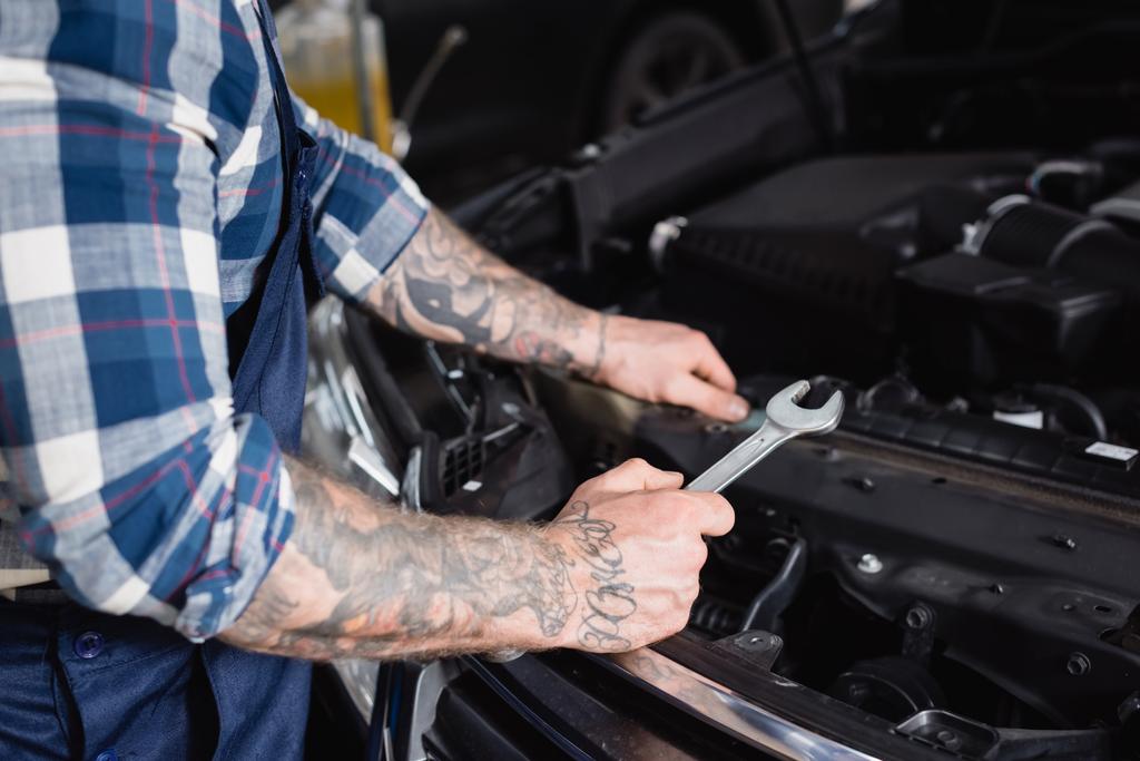 partial view of tattooed technician holding wrench near car engine compartment on blurred foreground - Photo, Image