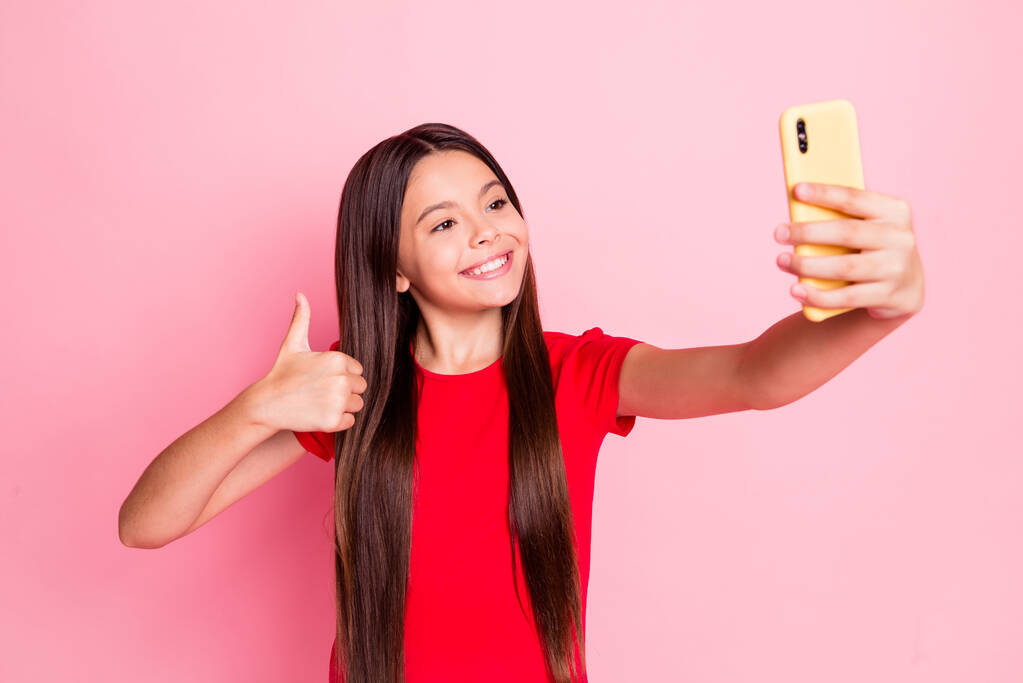 Fotografie roztomilé krásné malé dámy dlouhé vlasy zvýšit ruční držení telefonu show thumb-up, aby selfie zubaté zářící úsměv oblečení ležérní stylové červené tričko izolované růžové barvy pozadí - Fotografie, Obrázek