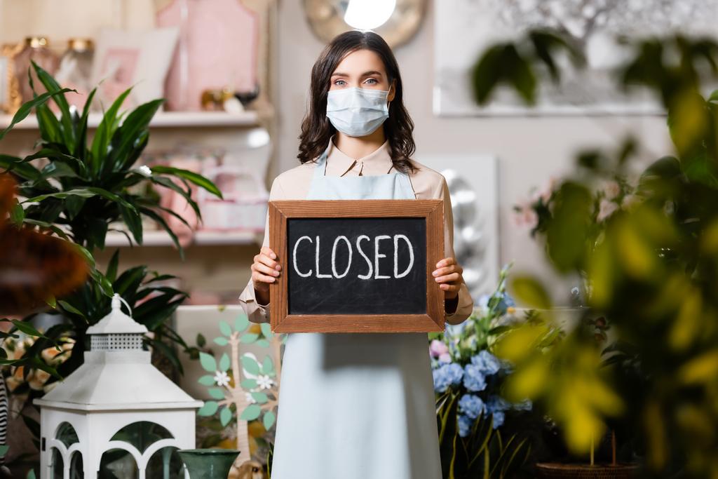 young florist in medical mask holding board with closed lettering near plants on blurred foreground - Photo, Image