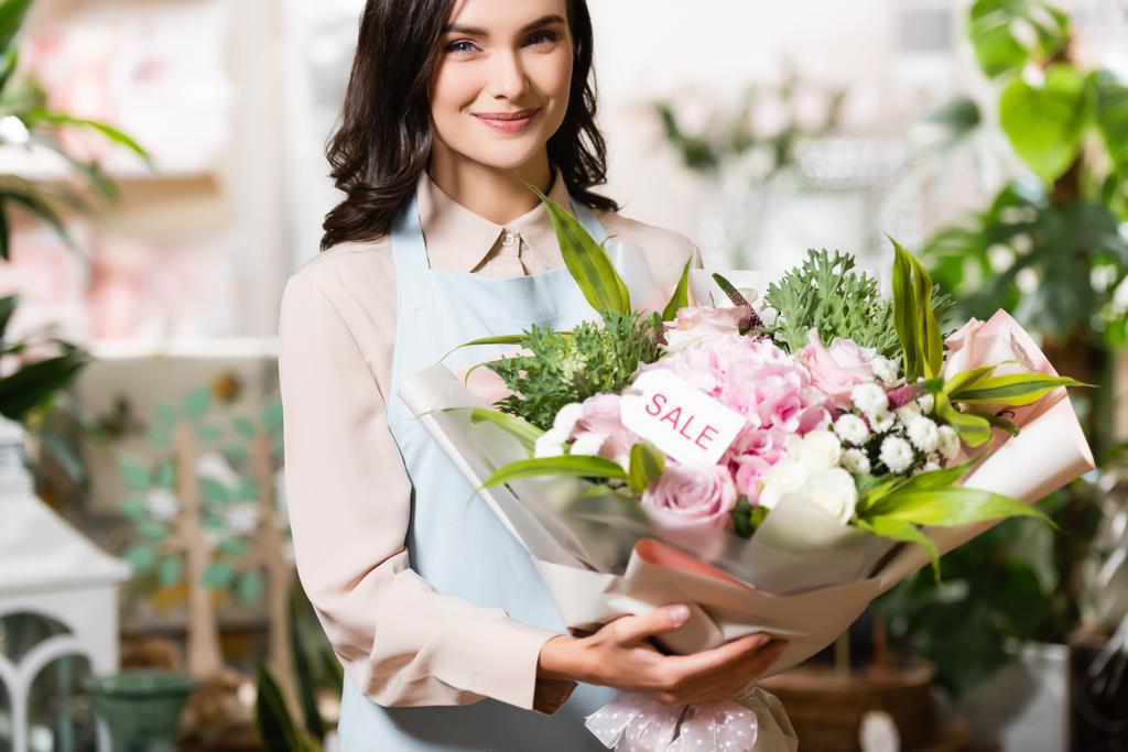 gelukkig bloemist bedrijf boeket met verkoop belettering op tag, terwijl het kijken naar camera in de buurt van wazig planten op de achtergrond - Foto, afbeelding